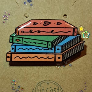 Pode incluir: Um alfinete de esmalte colorido com uma pilha de livros em laranja, verde, azul e laranja. O alfinete está em um cartão marrom com o texto "Outcast Charms" e decorações holográficas de estrelas e folhas.