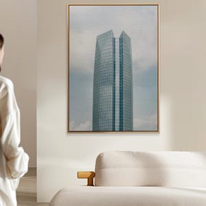 May include: A framed vertical wall art print featuring a tall, modern skyscraper with a glass facade against a cloudy sky. The building has a distinct angular peak and a central vertical column, rendered with a textured, painterly finish.