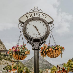 May include: A decorative street clock with the word "FRISCO" at the top. The clock face is white with black numerals and hands. Two hanging baskets filled with colorful flowers are suspended below the clock. The clock reads approximately 4:55.