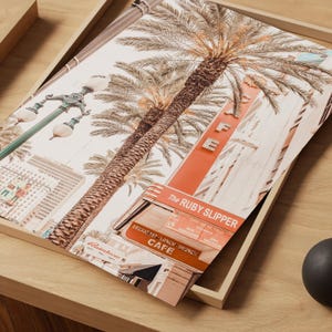 May include: A framed print featuring a vintage-style image of a cafe with the text "The Ruby Slipper Cafe". The print showcases palm trees, a street lamp, and buildings in a sepia-toned color palette. The print is displayed on a wooden tray.