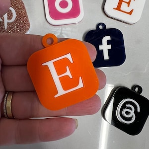 May include: A collection of colorful acrylic keychains featuring social media logos and letters. The keychains include an orange square with a white "E", a pink Instagram logo, a white square with an orange "E", a navy blue Facebook logo, and a black at symbol.
