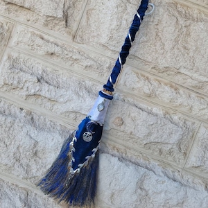 May include: A decorative broom featuring a long handle wrapped in dark blue velvet and silver ribbon. The base is adorned with white and blue velvet fabric, a silver crescent moon charm, a pentacle charm, and blue-dyed natural straw bristles.