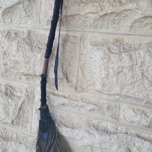 May include: A handcrafted broom with a dark handle and a black, decorative head. The broom's bristles are a mix of brown and tan. A black ribbon is wrapped around the handle and a black strap hangs from the top.
