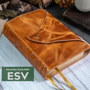 May include: A brown leather-bound MacArthur Study Bible, ESV edition, with a flap closure secured by a brass button. The book has gold page markers and is set on a light wood-grain surface. The cover has a distressed leather texture.