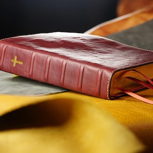 May include: A burgundy leather-bound book with a gold cross emblem. The book has a textured surface and visible page edges. Two red ribbon bookmarks extend from the pages. The book rests on a yellow and gray surface.
