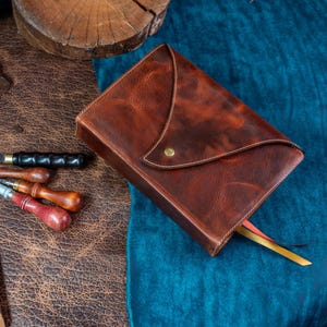 May include: A handcrafted brown leather journal with a unique flap closure and a brass stud. The journal has a textured appearance and a ribbon bookmark. Leatherworking tools, including a knife and awls, are arranged nearby.