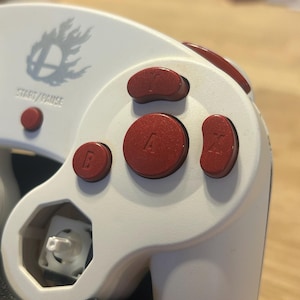 May include: A white video game controller featuring metallic red action buttons labeled A, B, X, and Y. The controller includes a small red Start/Pause button and a grey logo depicting a sphere with flames. One analog stick is missing, revealing the white internal mechanism.