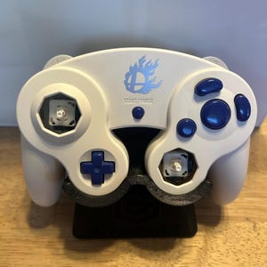 May include: A white video game controller featuring blue buttons, a blue D-pad, and a blue Smash Bros logo above the Start/Pause button. The controller has two exposed analogue stick sockets without the sticks attached and rests on a black stand.