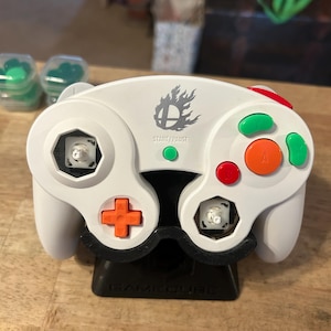 May include: A white video game controller with missing analog sticks, featuring an orange D-pad, a large orange A button, a red B button, and two green buttons. The center displays a grey flame logo above the text START/PAUSE and a green button.