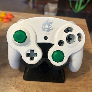 May include: A white video game controller featuring two green analogue sticks, a D-pad, and action buttons. The centre displays a flaming Smash Bros logo above the text START/PAUSE. The controller rests on a black stand embossed with the word GAMECUBE.