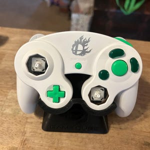May include: A white video game controller featuring a Smash Bros logo and the text START/PAUSE. The controller has bright green buttons and a D-pad, with two analog stick caps removed. It is mounted on a black stand with the word GAMECUBE embossed.