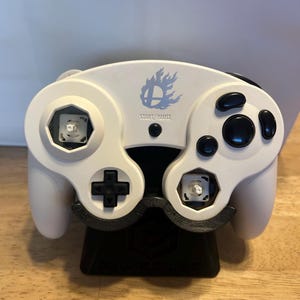 May include: A white video game controller with black buttons and a black D-pad. The controller features a blue flame logo above the text START/PAUSE. The two main analogue stick caps are missing, revealing the white internal mechanical stick boxes.