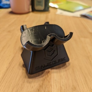 May include: A black 3D-printed controller stand featuring a contoured cradle top and a tapered base. The front of the base displays the embossed GameCube logo and the text GAMECUBE in capital letters, showcasing a layered texture finish.