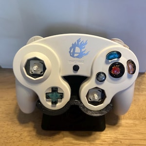 May include: A white video game controller featuring a blue flame logo and the text START/PAUSE. The controller has two exposed analog stick bases, a teal D-pad, and four colorful, textured action buttons in blue, red, and orange hues.