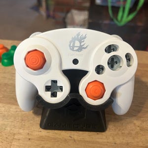 May include: A white video game controller with orange analogue sticks and a black base stand. The controller features a grey Smash Bros logo above the Start/Pause button. The base stand is black and has the word GAMECUBE embossed on the front surface.