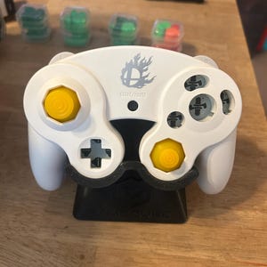 May include: A white video game controller featuring two yellow analogue sticks, a black D-pad, and a grey Super Smash Bros. logo above the Start/Pause button. The controller is resting on a black plastic stand with the word GAMECUBE embossed on the base.