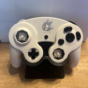 May include: A white video game controller featuring a black D-pad, black buttons, and two exposed mechanical switches where the analogue sticks are typically located. The centre displays a grey flame logo and the text START/PAUSE above a black button.