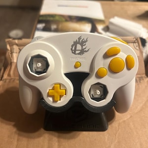 May include: A white video game controller with yellow buttons and a yellow directional pad. The controller features a Super Smash Bros logo above the Start/Pause button. Two analog stick caps are missing, revealing the internal mechanisms.