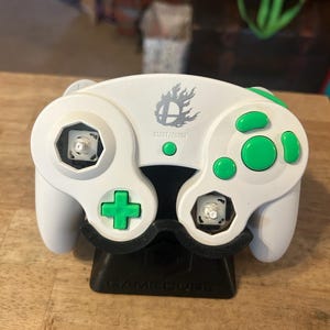 May include: A white video game controller with bright green buttons and a gray Smash Bros logo above the Start/Pause button. The controller features two exposed analog stick bases without caps and rests on a black GameCube branded display stand.