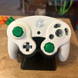 May include: A white video game controller featuring a Smash Bros logo and the text START/PAUSE, equipped with two green thumbsticks and a black D-pad. The controller rests on a black stand with the embossed text GAMECUBE on the front base.