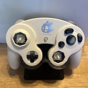 May include: A white video game controller featuring a blue flame logo above the text START/PAUSE. The controller has black buttons, a black directional pad, and two missing analogue stick caps, revealing the white internal mechanical components.