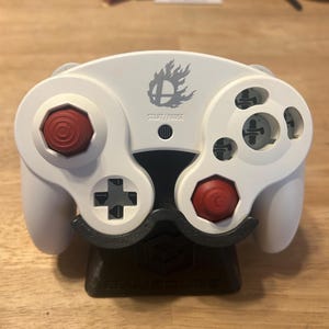 May include: A white video game controller featuring a gray Super Smash Bros. flame logo above the START/PAUSE text. The controller has two red analog sticks, a gray directional pad, and gray action buttons, all resting on a black display stand.