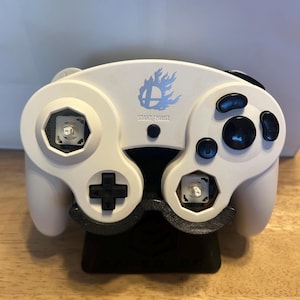 May include: A white video game controller featuring a blue flame logo above the text START/PAUSE. The controller has black buttons, a black directional pad, and two exposed white analog stick bases without the thumb caps attached.