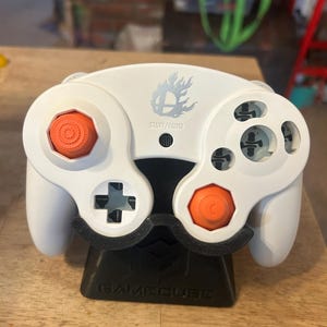May include: A white video game controller featuring orange analog sticks, a black D-pad, and a Super Smash Bros. logo with the text START/PAUSE printed above a small black button. The controller is resting on a black stand labeled GAMECUBE.