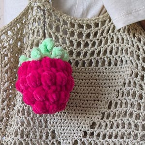 May include: A beige crocheted tote bag with a large, bright pink strawberry charm. The strawberry has green leaves. The bag has a large open weave design. The bag is being held by a person.