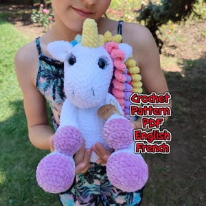 May include: A white plush unicorn toy with a yellow horn, long eyelashes, and a multi-colored mane featuring pink, yellow, and blue curls. The unicorn has light purple feet and a small brown tag. Text reads: Crochet Pattern PDF English French.