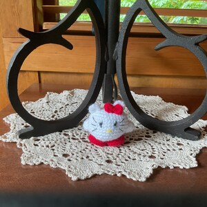May include: A white crocheted Hello Kitty character with red bow and red shoes sitting on a white crocheted doily.