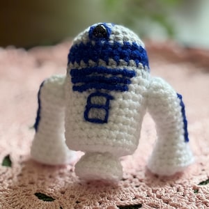 May include: A handmade crochet figure of R2-D2, the iconic droid. The figure is primarily white with blue and gray accents on the head and details. It has a small, rounded body with two arms and a single foot.