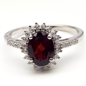 Puede incluir: Un anillo de plata con una gran piedra roja ovalada rodeada por un halo de piedras blancas más pequeñas.