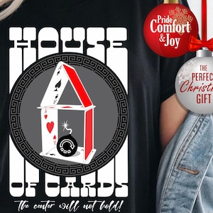May include: Black t-shirt with a graphic of a house of cards, featuring playing card suits and a bomb. The text "HOUSE OF CARDS" is above the graphic, with "the center will not hold!" below. Additional text includes "T-SHIRTS HOODIES TRANSFERS".