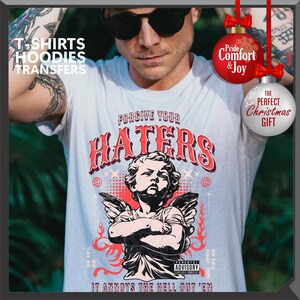 May include: White t-shirt with a graphic featuring the text "FORGIVE YOUR HATERS" above an illustration of an angel with wings. The shirt also includes the text "IT ANNOYS THE HELL OUT 'EM" and a parental advisory warning.
