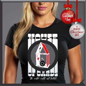 May include: Black t-shirt with a graphic of a house made of playing cards, with the text "HOUSE OF CARDS" and "The center will not hold!". The design includes a red and white color scheme, with a circular border. Christmas ornaments are in the background.