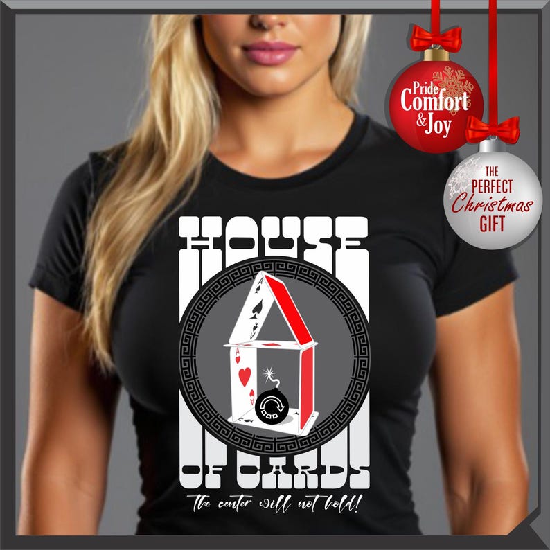 May include: Black t-shirt with a graphic of a house made of playing cards, with the text "HOUSE OF CARDS" and "The center will not hold!". The design includes a red and white color scheme, with a circular border. Christmas ornaments are in the background.