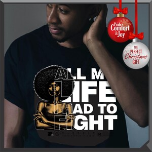 Może przedstawiać: Czarny t-shirt z grafiką kobiety z afro i napisem "ALL MY LIFE HAD TO FIGHT". Projekt jest w kolorach białym i złotym. Widoczna jest bombka świąteczna.