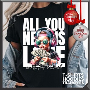 Puede incluir: Camiseta negra con el texto "ALL YOU NEED IS LOVE" y un gr&aacute;fico de un ni&ntilde;o con auriculares y gafas de sol, sosteniendo dinero. La frase "well, that's cash" est&aacute; escrita debajo. Incluye adornos navide&ntilde;os.