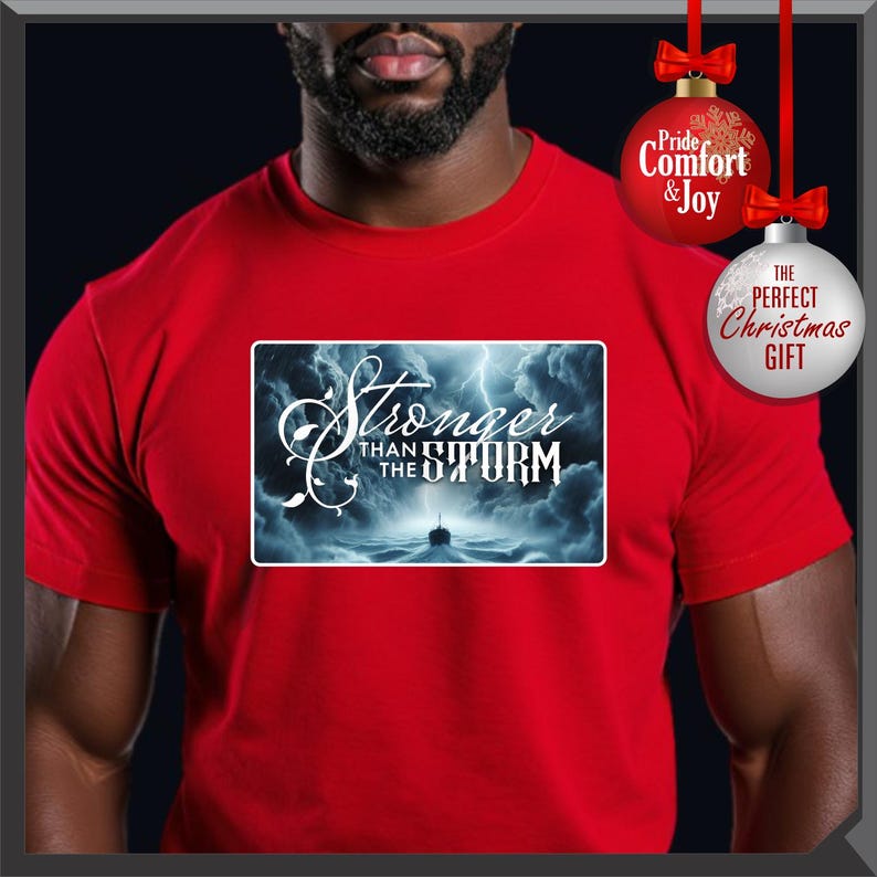 K&ouml;nnte beinhalten: Ein rotes T-Shirt mit einem rechteckigen Aufdruck mit dem Text "Stronger Than The Storm" in wei&szlig;er Schrift. Die Grafik zeigt ein Schiff auf See w&auml;hrend eines Sturms. Zwei Weihnachtskugeln h&auml;ngen im Hintergrund.