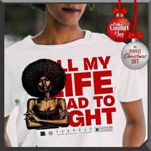 Może przedstawiać: Biała koszulka z grafiką kobiety z afro i napisem "ALL MY LIFE HAD TO FIGHT" w kolorze czerwonym. Koszulka zawiera r&oacute;wnież napis "INNER WARRIOR UNLEASHED". Ozdoby świąteczne w tle.
