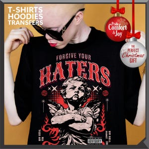 May include: Black t-shirt with the text "FORGIVE YOUR HATERS" in red, featuring an angel graphic. The shirt also has the text "T-SHIRTS HOODIES TRANSFERS" and "ADVISORY EXPLICIT CONTENT". Decorative Christmas ornaments are also visible.