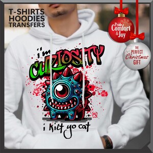 Może przedstawiać: Biała bluza z kapturem z grafiką z kresk&oacute;wkowym potworem z napisem "I'm Curiosity" i "i kilt yo cat". Projekt wykorzystuje kolory czerwony, niebieski i zielony. Bluza ma kaptur ze sznurkiem i długie rękawy. Widoczne są r&oacute;wnież ozdoby świąteczne.
