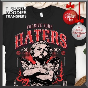 May include: Black t-shirt with a graphic featuring an angel and the text "FORGIVE YOUR HATERS." The design includes red accents and decorative elements. The shirt also has the text "T-SHIRTS HOODIES TRANSFERS."