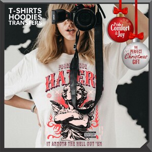 May include: White t-shirt with a graphic of an angel and the text "FORGI OUR HATER" and "IT ANNOYS THE HELL OUT 'EM". The image also includes text that says "T-SHIRTS HOODIES TRANSFERS".