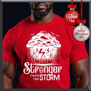 K&ouml;nnte beinhalten: Rotes T-Shirt mit wei&szlig;er Grafik eines Gewitterwolken, Blitzen und dem Text "Stronger Than The Storm". Das Shirt hat auch zwei Weihnachtsbaumschmuck-Dekorationen mit Text. Ein bequemes T-Shirt.