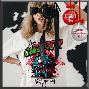 Może przedstawiać: Biała koszulka z kolorową grafiką potwora i napisem "I'm CURIOUS i kilt yo cat". Potw&oacute;r jest niebieski, czerwony i zielony z dużym okiem i kolczastymi detalami. Koszulka ma okrągły dekolt.