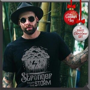 K&ouml;nnte beinhalten: Schwarzes T-Shirt mit einer grauen Grafik einer Gewitterwolke, Regen und Blitzen. Der Text "Stronger Than The Storm" befindet sich darunter. Das Bild zeigt auch eine Person mit schwarzem Hut und Sonnenbrille.