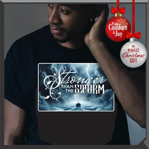 K&ouml;nnte beinhalten: Schwarzes T-Shirt mit dem Text "Stronger Than The Storm" in wei&szlig;er Schrift &uuml;ber einer st&uuml;rmischen Meereslandschaft. Das Design zeigt ein Schiff, das unter einem blitzgef&uuml;llten Himmel durch die Wellen segelt. Weihnachtsschmuck ist ebenfalls zu sehen.