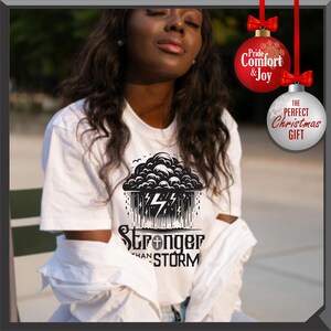 K&ouml;nnte beinhalten: Wei&szlig;es T-Shirt mit einem schwarz-wei&szlig;en Grafikdesign einer Gewitterwolke mit Blitzen und dem Text "Stronger Than Storm". Das Shirt hat einen Rundhalsausschnitt und kurze &Auml;rmel. Das Bild enth&auml;lt auch Weihnachtsschmuck.